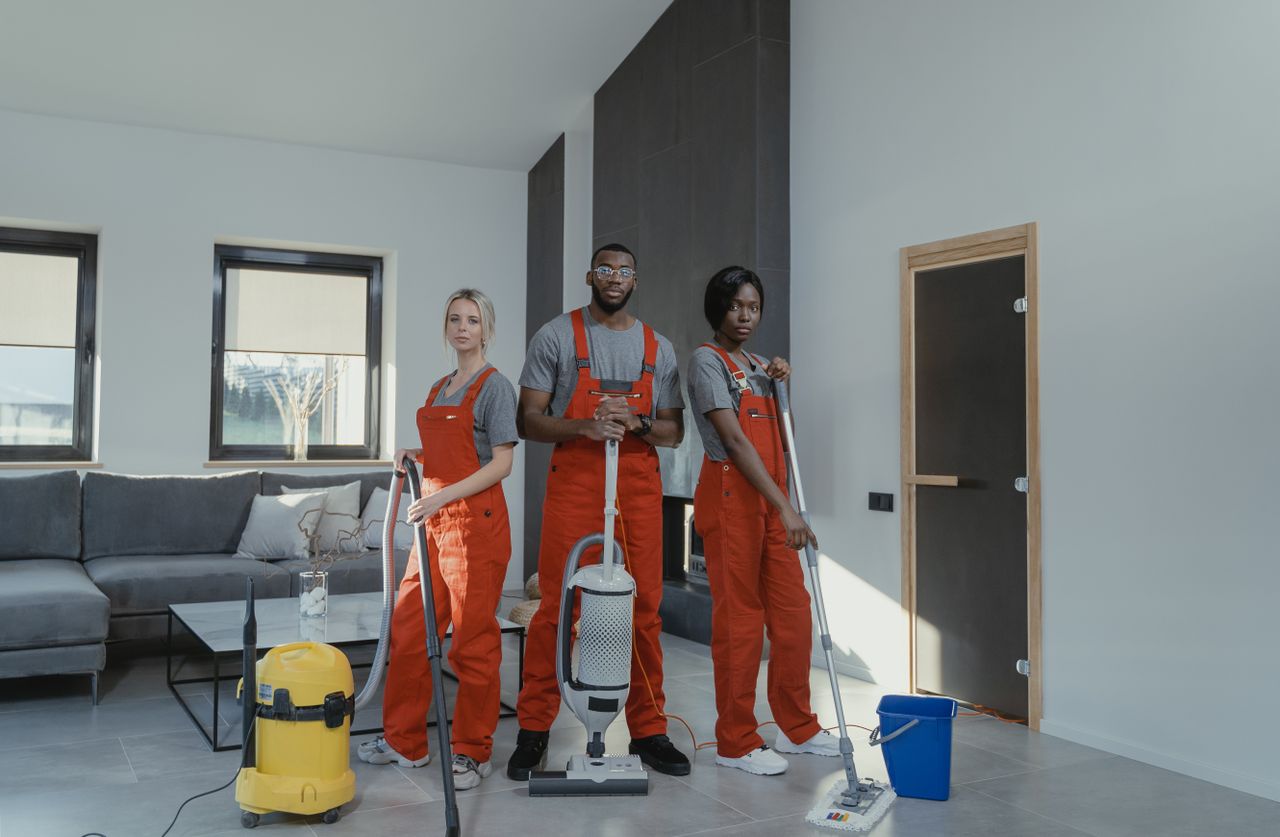 about-me Diverse cleaning team in red uniforms ready to sanitize a modern living room with vacuum and mop.
